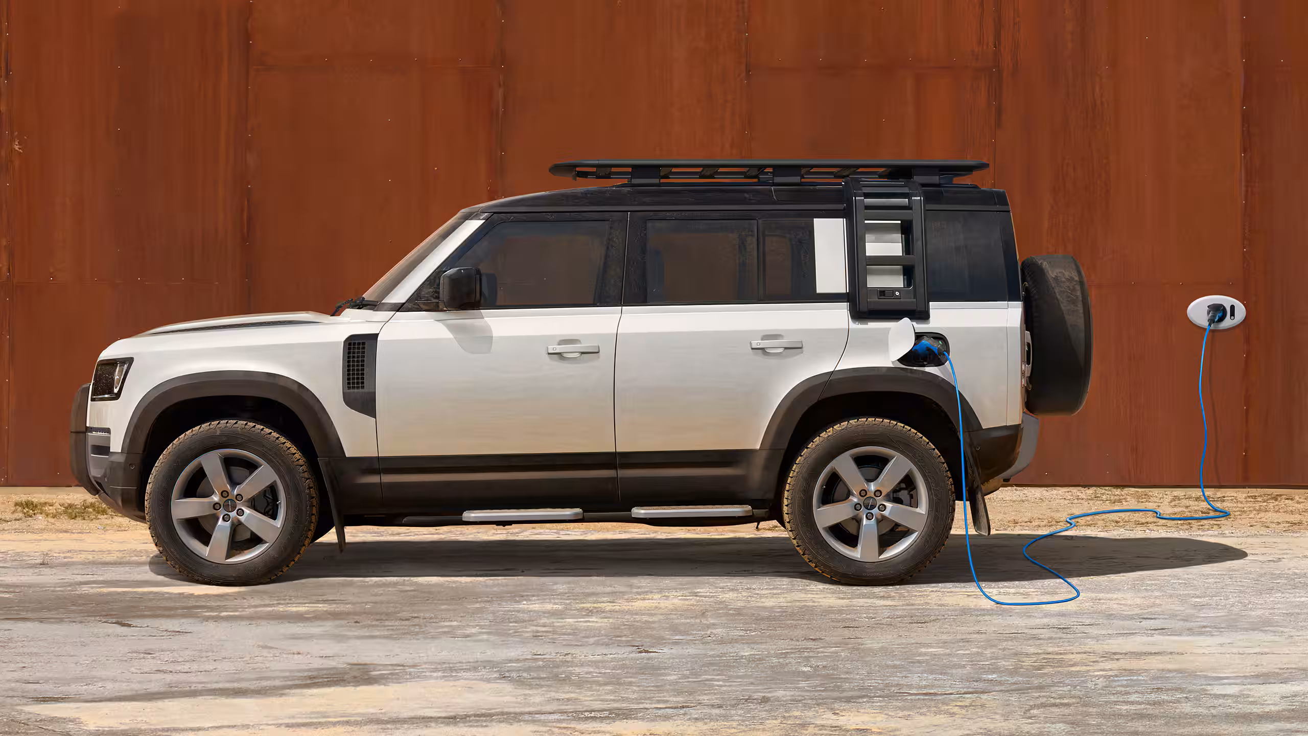 Vue de profil d’un Land Rover Defender blanc branché à une borne de recharge électrique, stationné devant un mur en métal brun.