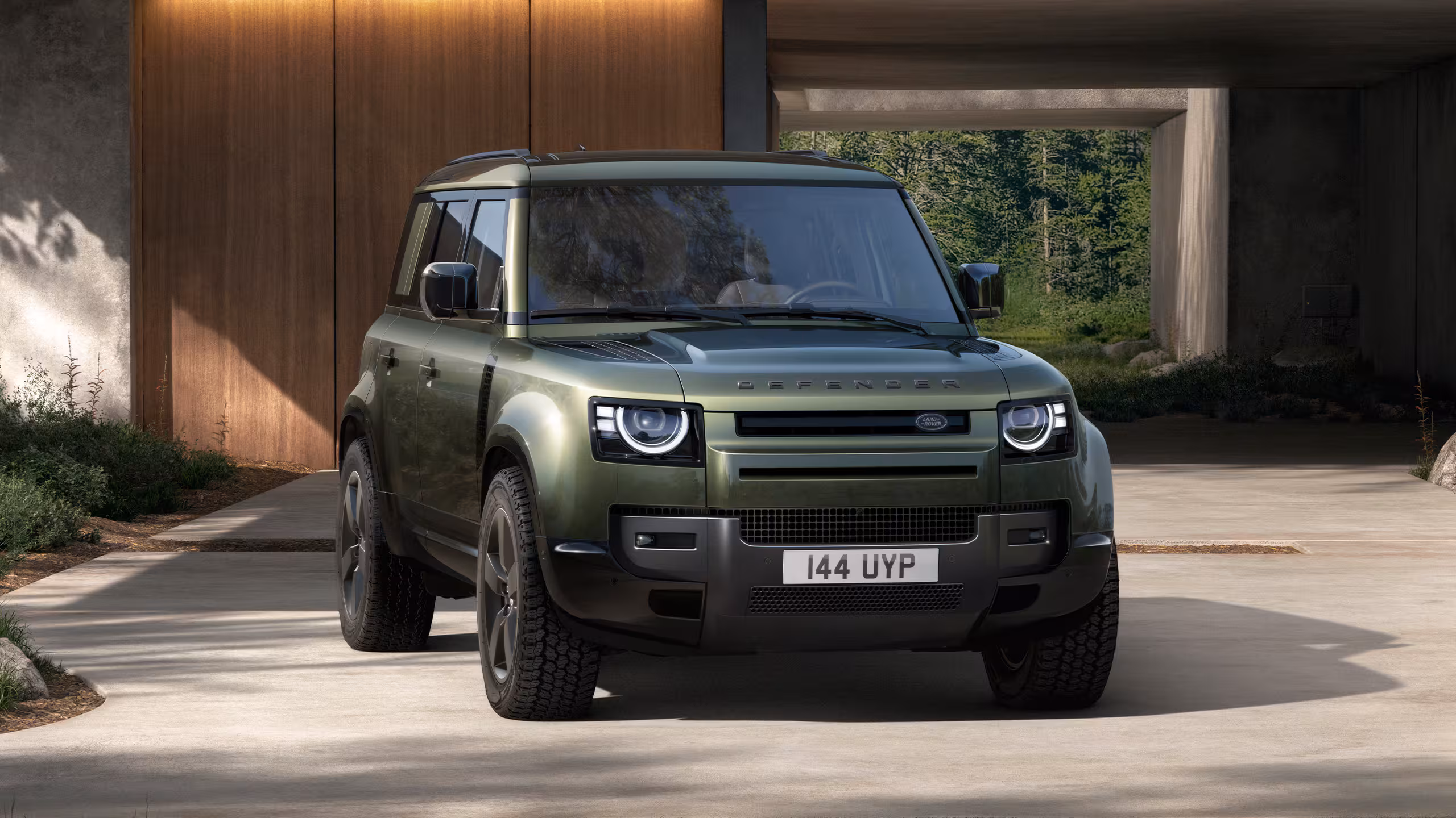 Land Rover Defender vert vu de face, stationné devant une maison moderne avec façade en bois et ouverture sur un espace extérieur.